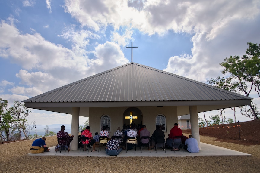 People sit on chairs under a small hilltop chapel with a cross on its roof, overlooking a rural landscape.