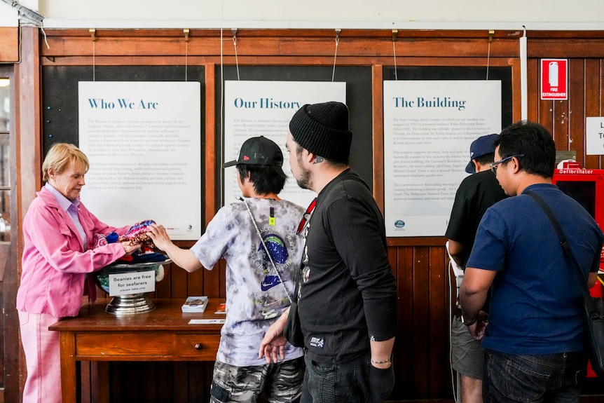 A woman dressed in pink stands at a wooden table and hands four men knitted beanies.