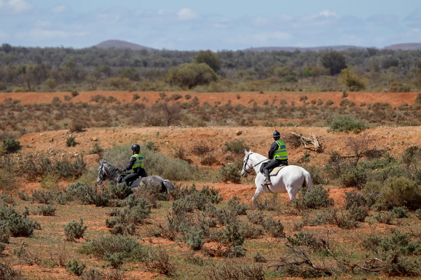 Two horses are ridden by police officers wearing uniform and fluoro vests. They walk along rugged outback terrain.