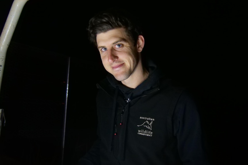 A man wearing a black hoodie in the darkness smiles at the camera.