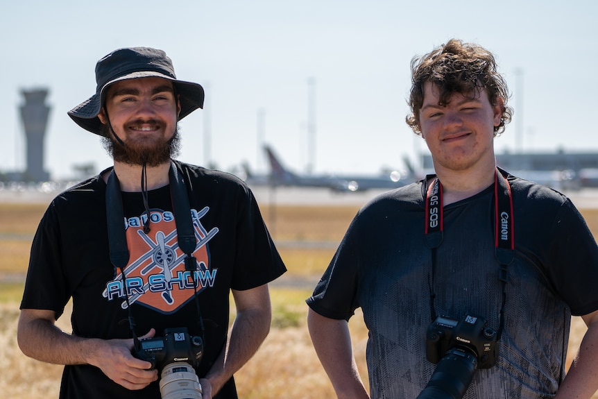 Two men with camera straps in front of an airport tower and aeroplanes