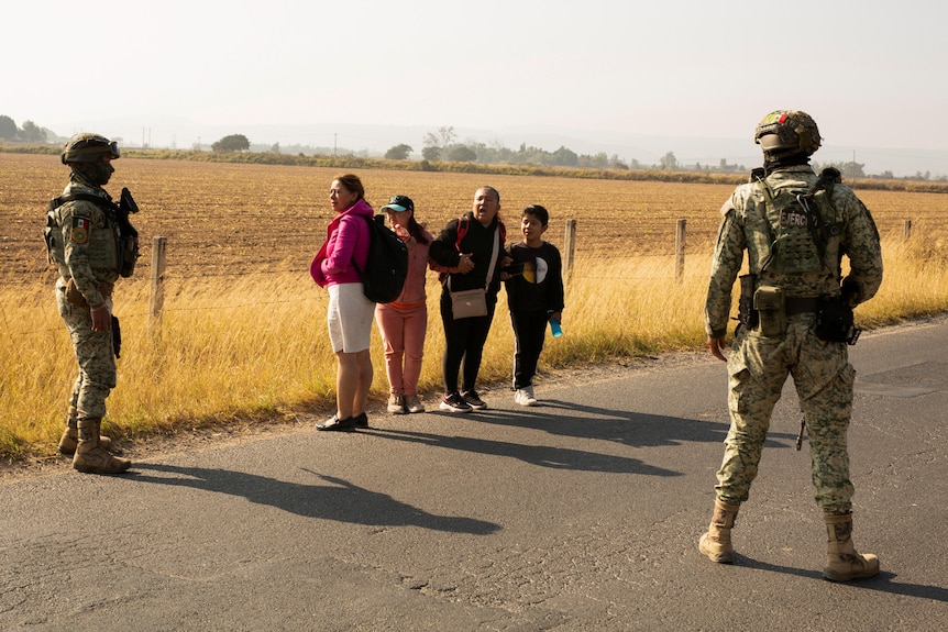 Four bypassers are stopped by two military workers on a road.