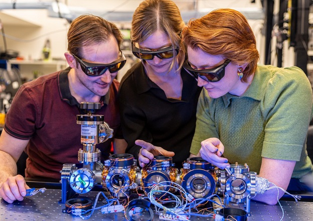 Three people wearing protective glasses, standing next to equipment associated with optical atomic clocks