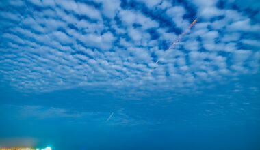 An orange and white streak cuts diagonally through a cloudy sky and down to Earth in the distance. The clouds have an unusual, rippling, wave-like pattern.