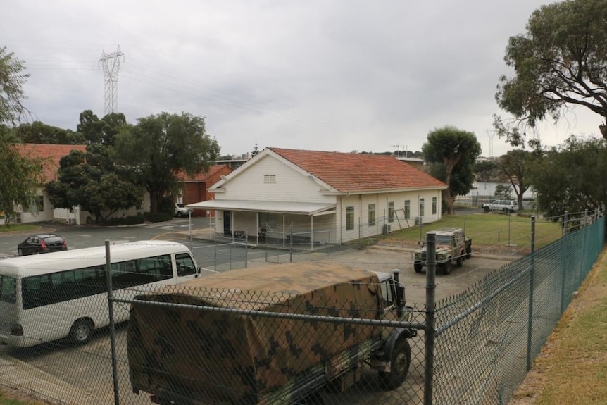 A shot of an army barracks building from hehind a fence with some army and civilian vehicles visible parked outside. 