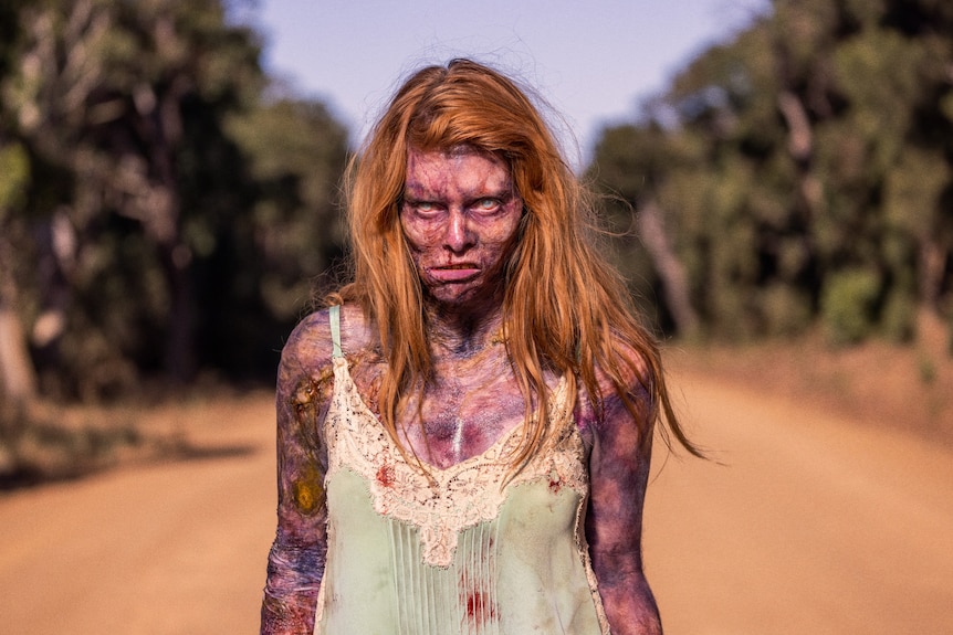A female zombie with long auburn hair and a menacing look on her face standing on a country road.