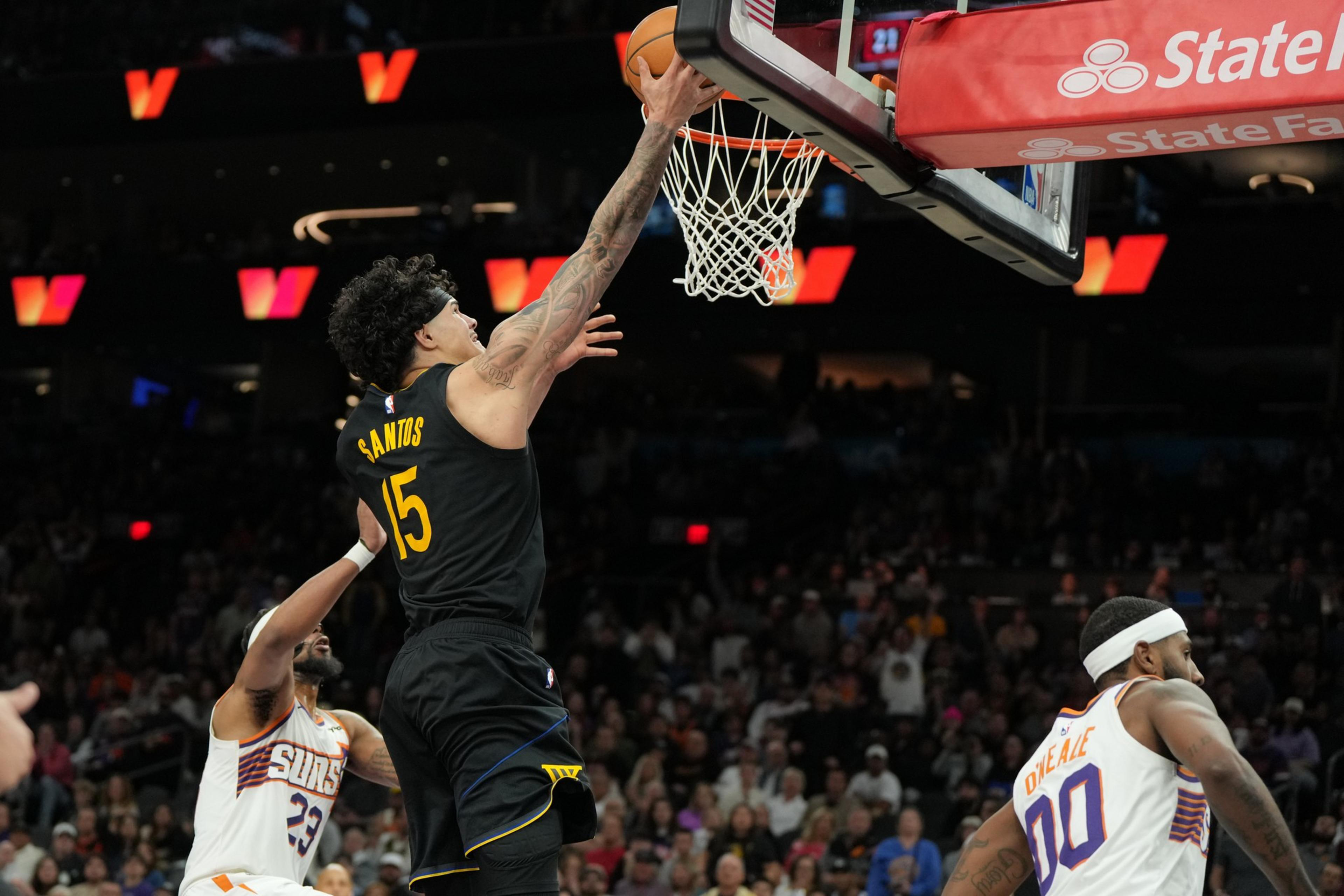 A basketball player in a black jersey labeled “Santos” leaps to score near the hoop while two opposing players in white jerseys watch nearby.