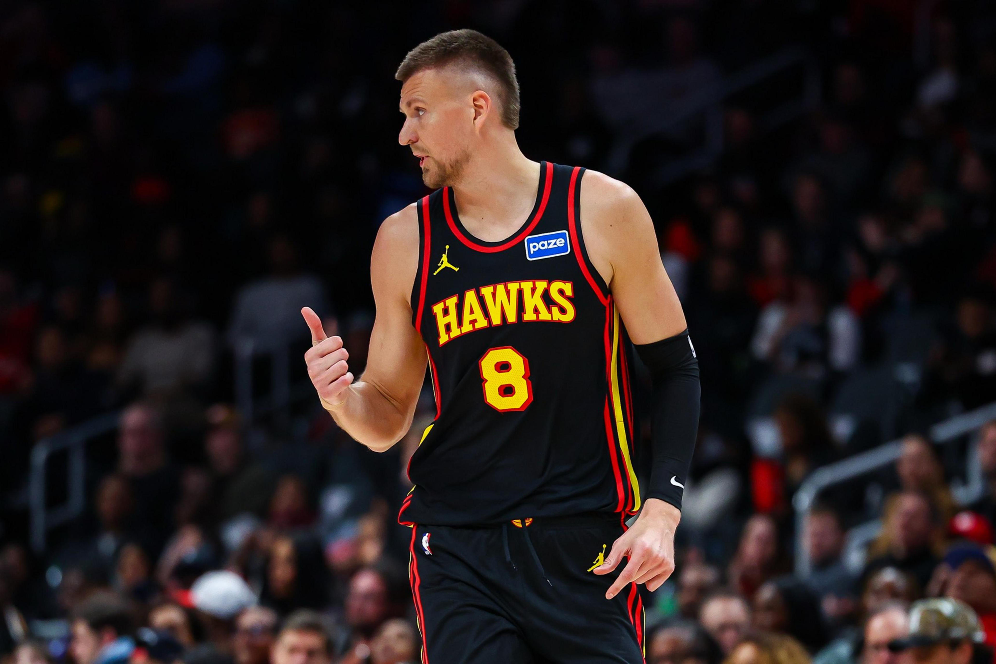 A basketball player wearing a black Hawks jersey with the number 8 gives a thumbs-up, looking to his left on a crowded court.