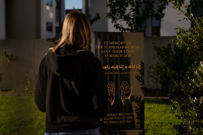 The back of a woman with blonde hair and a black jumper, standing at a memorial plaque with gold writing.