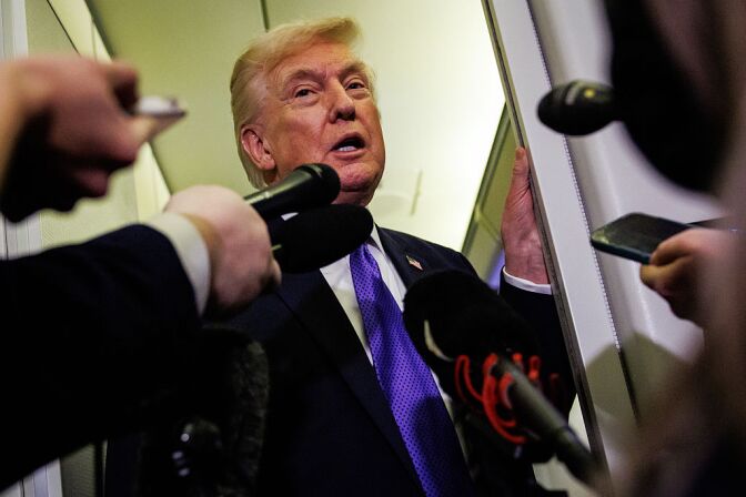 A low angle view of President Donald Trump, a man with light skin tone, wearing a dark blue suit and violet spotted tie, speaking to reporters holding and pointing microphones to him as he stands at a doorway on a plane.