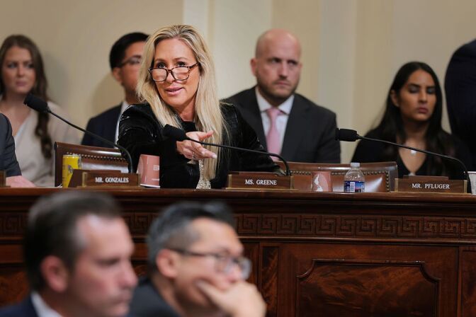 Rep. Marjorie Taylor Greene, a woman with light skin tone, blonde hair, wearing a black coat, speaks behind a wooden desk behind a nameplate that reads "Ms. Greene." There are more people, slightly out of focus, in the background and foreground.