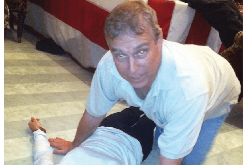 A caucasian man bent over a person laying on the floor, on all fours, as he looks at the camera