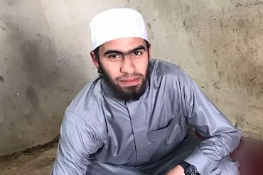 A man wearing a traditional Muslim cap and long shirt crouches on a concrete floor. He is looking at the camera.
