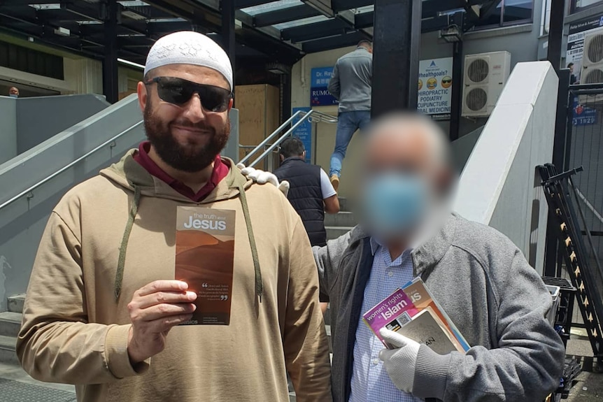 A man wearing traditional Muslim cap and shirt stands holding a pamphlet. A man whose face is blurred stands next to him.