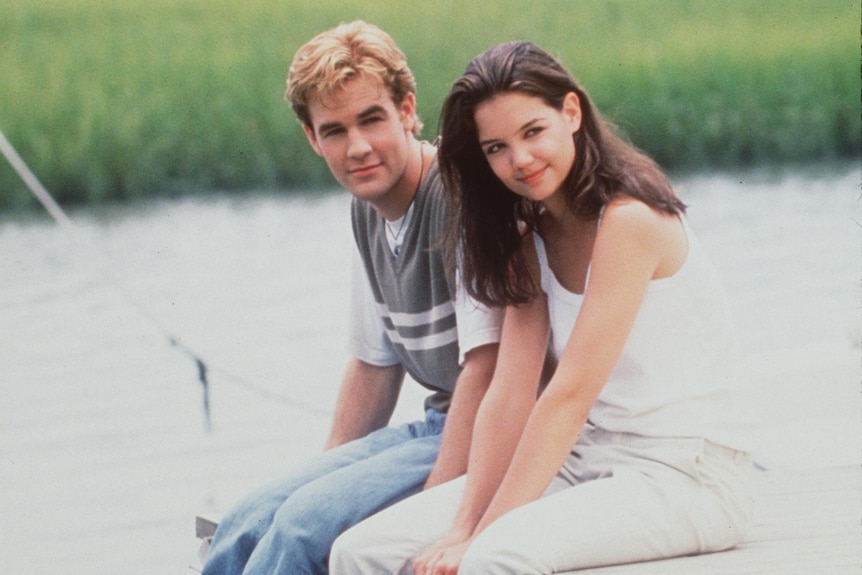 A man and woman sit on a dock with water behind them. 