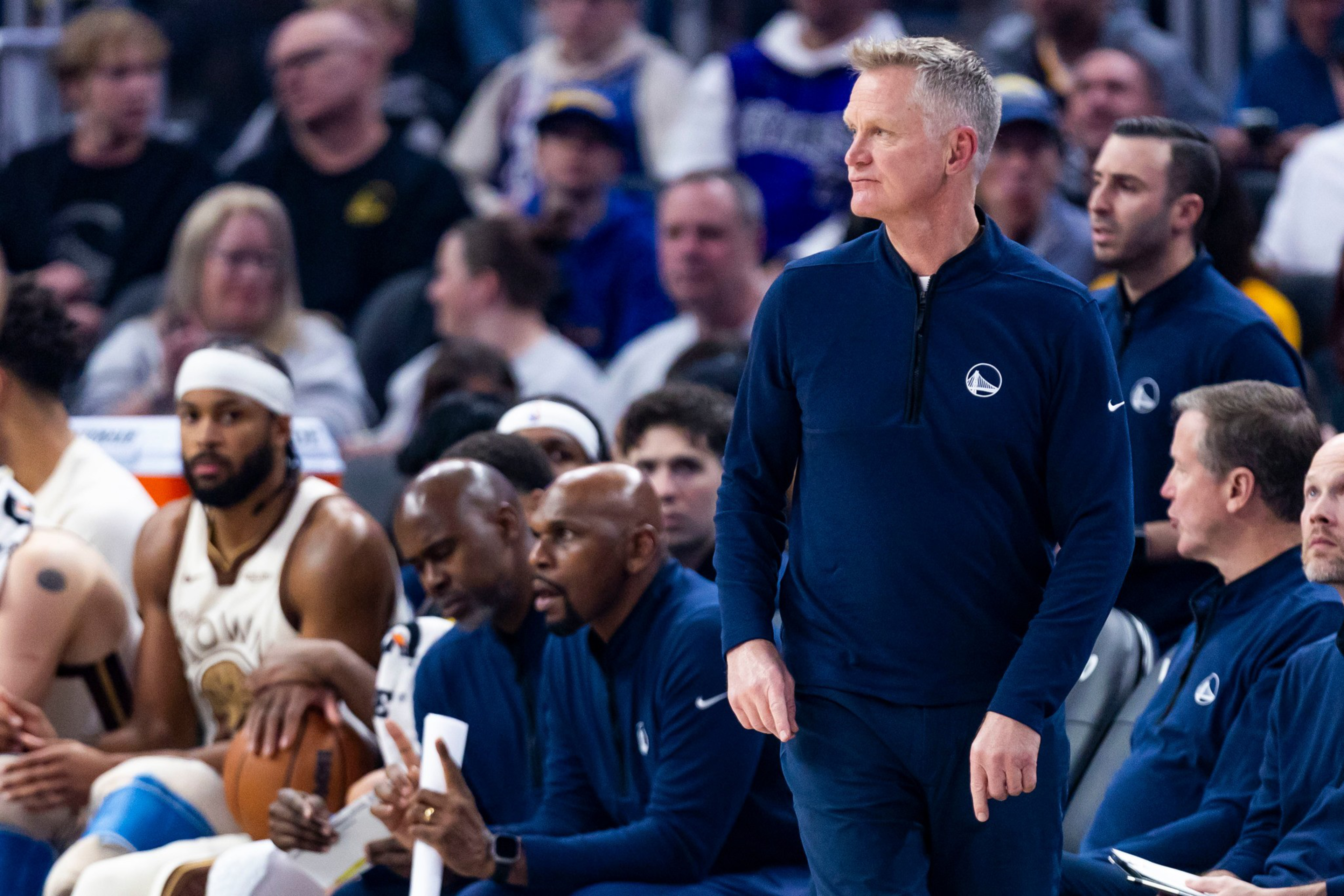 A basketball coach in a navy Golden State Warriors jacket stands focused near the bench while players and staff sit behind him during a game.
