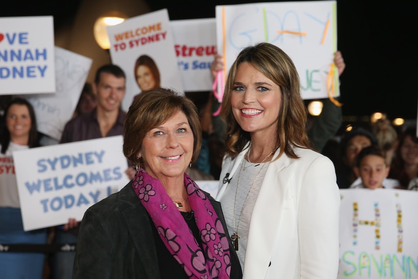 A photo of Savannah Guthrie alongside her mother Nancy behind a crowd with posters.