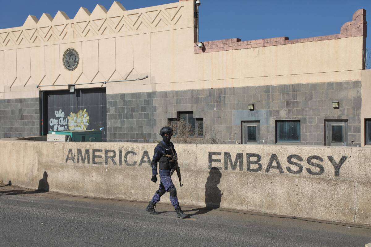 A soldier walks by a low wall with the words "American Embassy" on it.