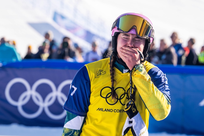 A woman in snowboarding gear puts her hand to her mouth in front of a crowd at an Olympic event.