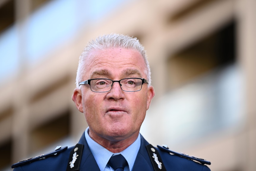 An up close photo of Mal Lanyon in his NSW Police suit uniform, black glasses, and white hair.