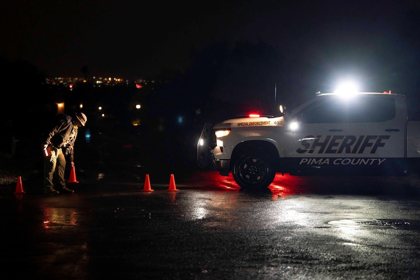 A sheriff places orange witches hats on a wet road at night near a sheriff's car