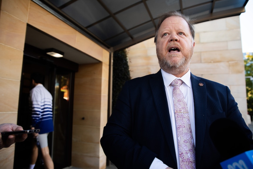 A man in a suit and tie, Matthew Swinbourn, speaking outside a building.
