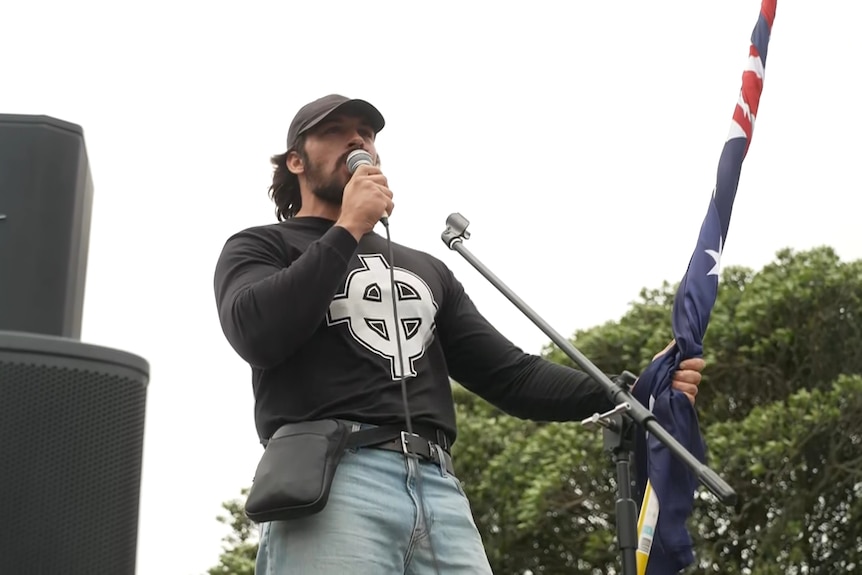 Brandan Koschel at the rally, wearing a black shirt with a Celtic cross, and holding an Australian flag and microphone.