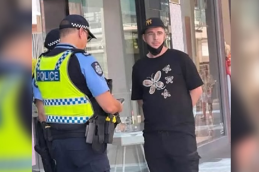 Police speak to a man wearing a black butterfly print T-shirt, baseball cap and face mask