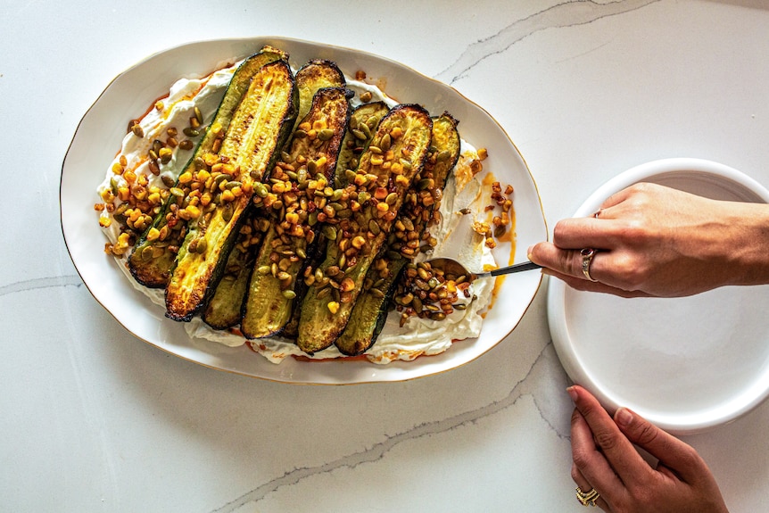 Baked zucchinis with corn and pumpkin seeds on top, sitting on a white spread on a platter.