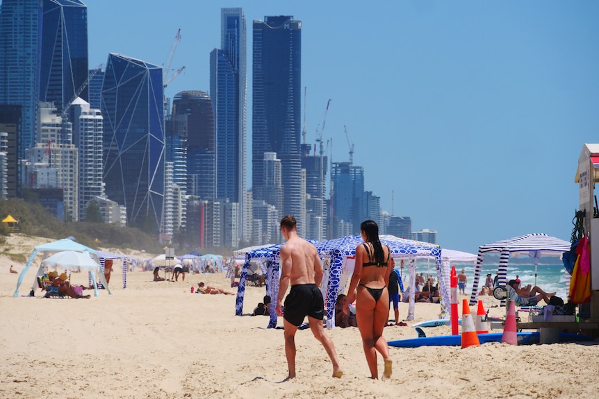 A man and woman walk away from the camera on a crowded beach by a city skyline.