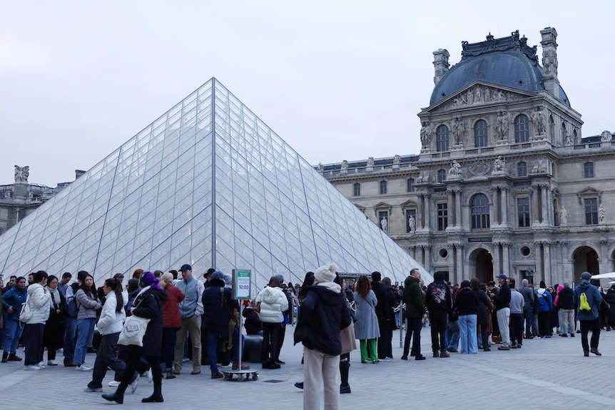 Queues of tourists lined up alongside the Louvre's glass pyramid and a stone building.