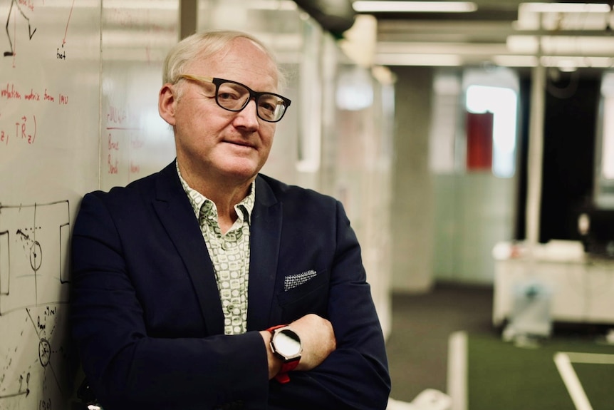 A silver-haired man in glasses and a dark blazer leans against whiteboard in a tutorial room.