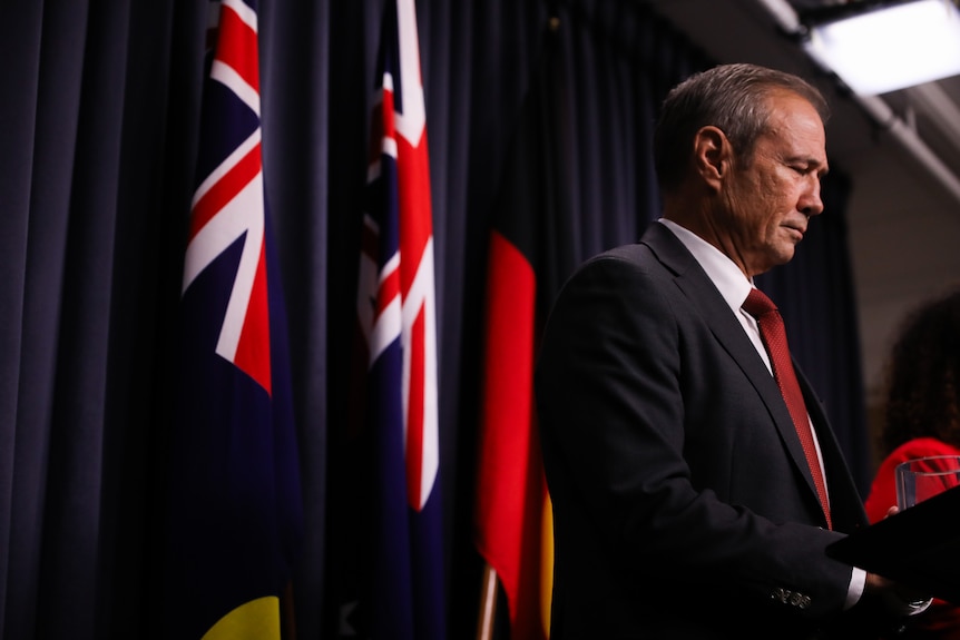 Roger Cook looks down in front of flags.