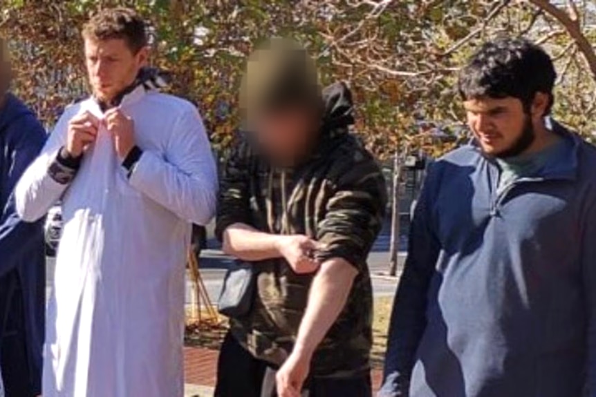 Four men stand in a row outdoors. One, wears a white traditional long shirt, another looks down.