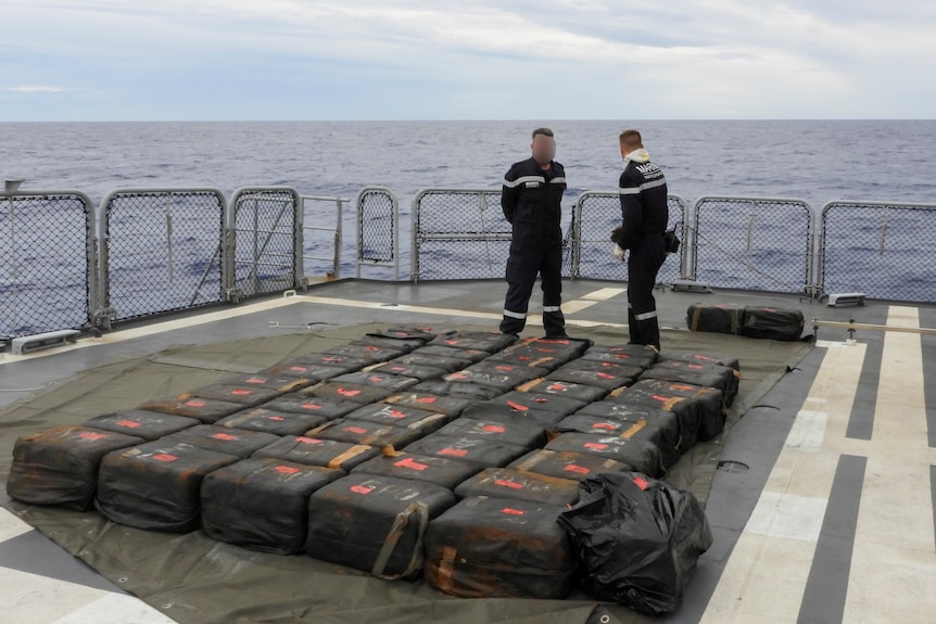 Two men stand on a ship next to many bags of cocaine.