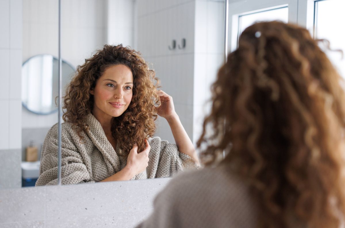 A mid adult woman with curly hair wearing a cozy beige robe looks thoughtfully at her reflection in a bright bathroom with white tiles and natural light.