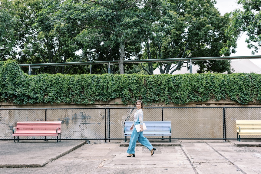 A smiling, dark-haired woman holds a binder as she walks down a concrete path near a park.