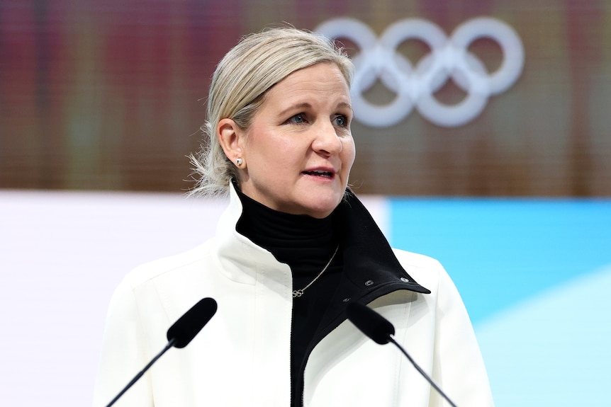 A blonde woman wearing a white jacket speaks at a podium in front of Olympic rings.