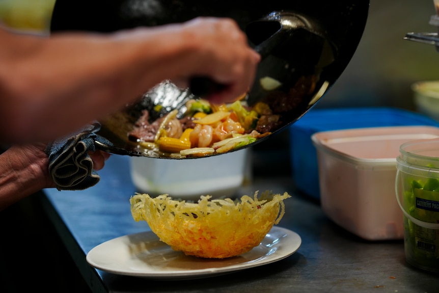 A pair of hands can be seen serving stir-fried ingredients from a wok to a plating dish.