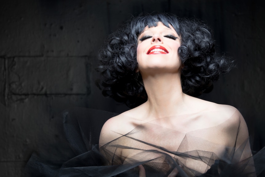 Meow Meow, dramatically made up with curly black hair and pale skin, grins with her eyes shut. Black background.