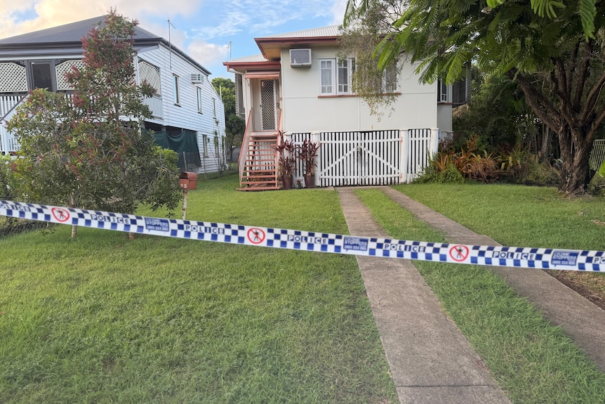 Police tape cordons off a light-coloured, Queenslander-style house with a large tree out the front.