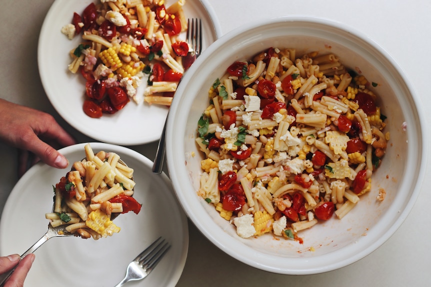 Close-up of hands serving pasta salad with corn, feta, tomatoes and basil on a plate, from a large bowl of pasta salad.