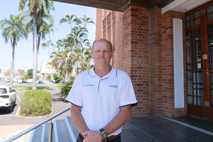 Middle aged man with semi balding hair, white shirt, arms crossed, trees, gardens and roads in background