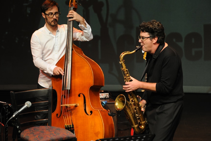 Two men performing, one is playing the cello and the other the saxophone. 