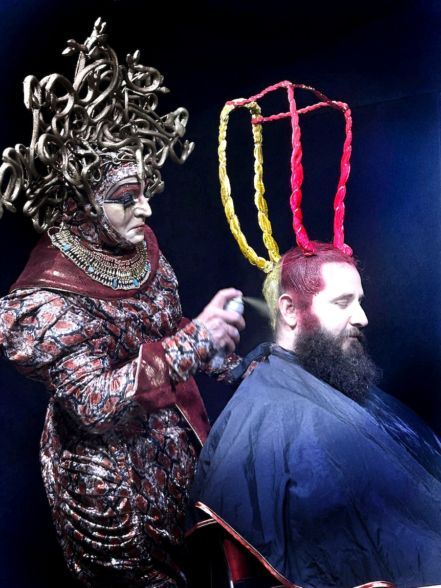 A person in strange outfit turns a person's hair into a strange sculpture