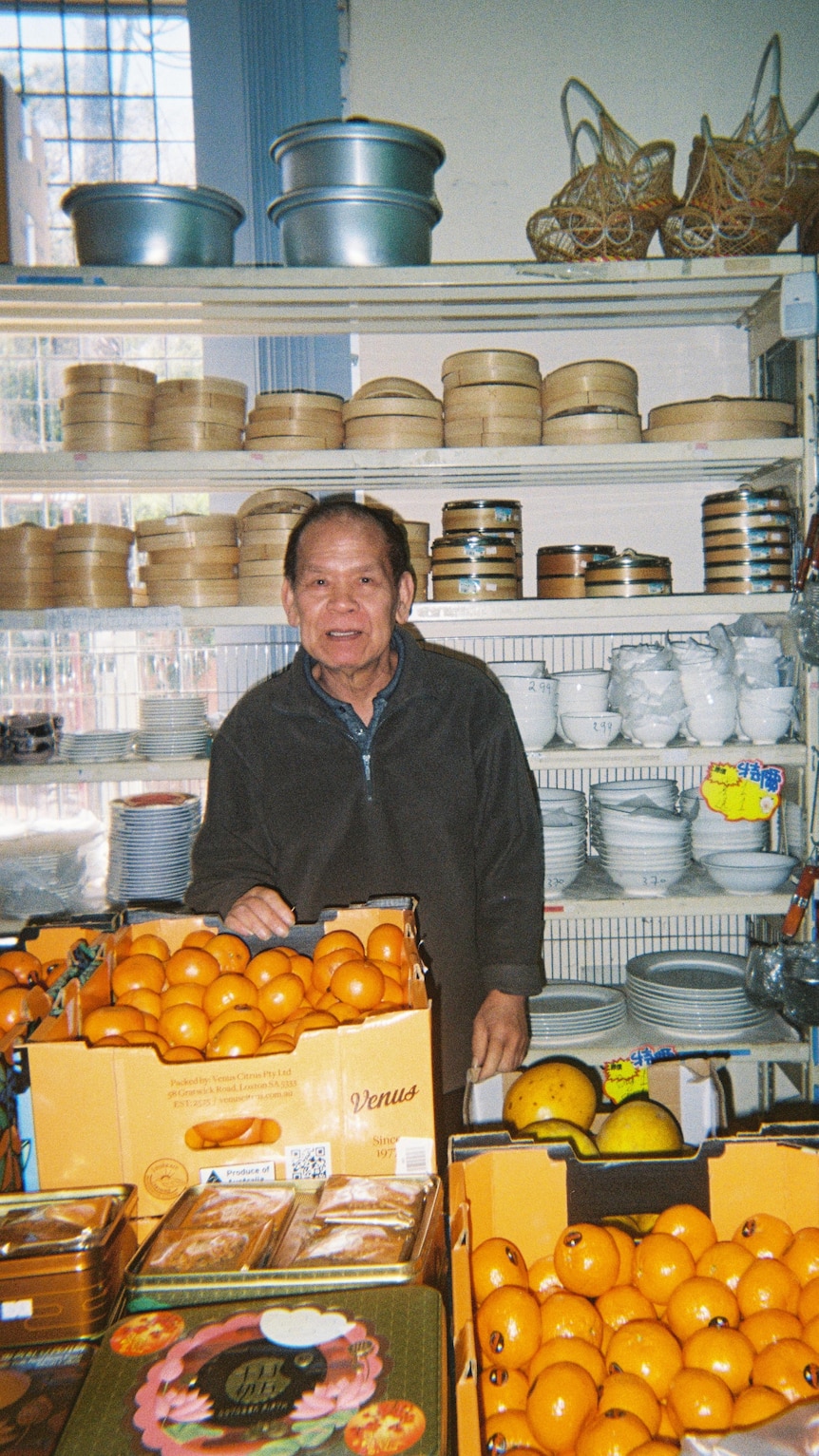 An asian man stands in front of shelves of bamboo steamers and behind boxes of orange citrus fruit.
