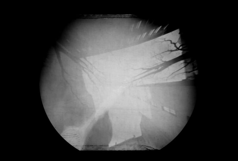 A blurred, black and white photo shows a circular frame with faint outlines of bare tree branches and the edges of buildings, creating a ghostly, abstract scene against a dark background.