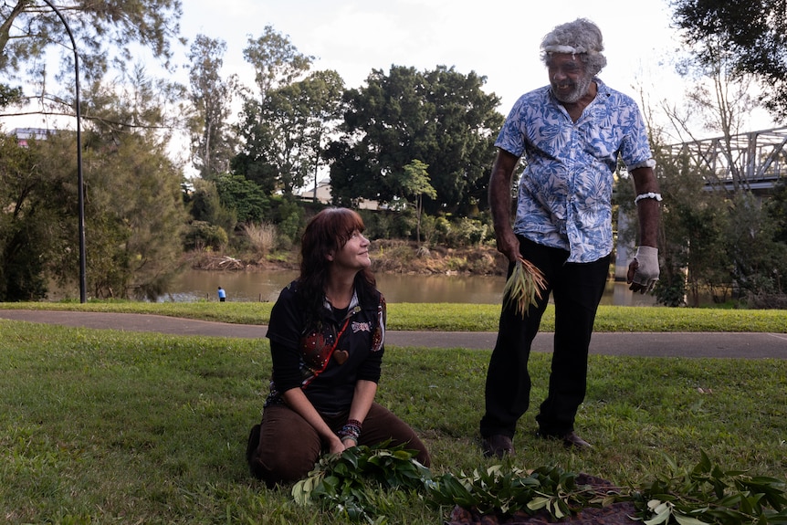 Carlie Atkinson, a middle-aged Aboriginal woman, kneels in a park, looking up an older Aboriginal man, holding foliage.