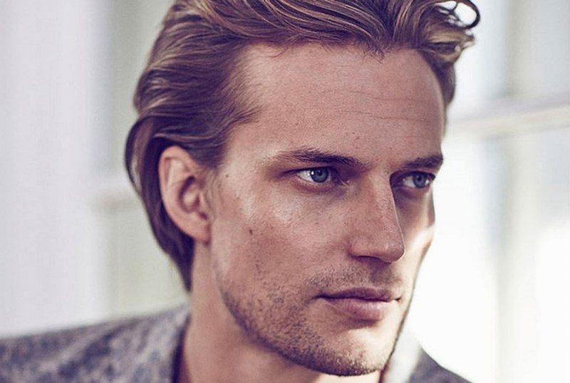 Close-up portrait of a young man with tousled blonde hair and striking blue eyes, showcasing a thoughtful expression and well-defined facial features against a soft, neutral background.
