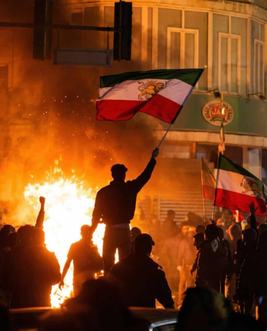Protesters wave Iranian flags as flames light up a nighttime street during unrest.